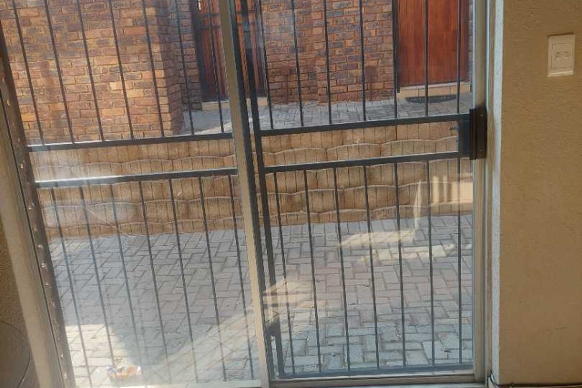 Interior view of a living area featuring ceramic floor tiles and a sliding glass door with a security gate leading to an exterior brick patio.
