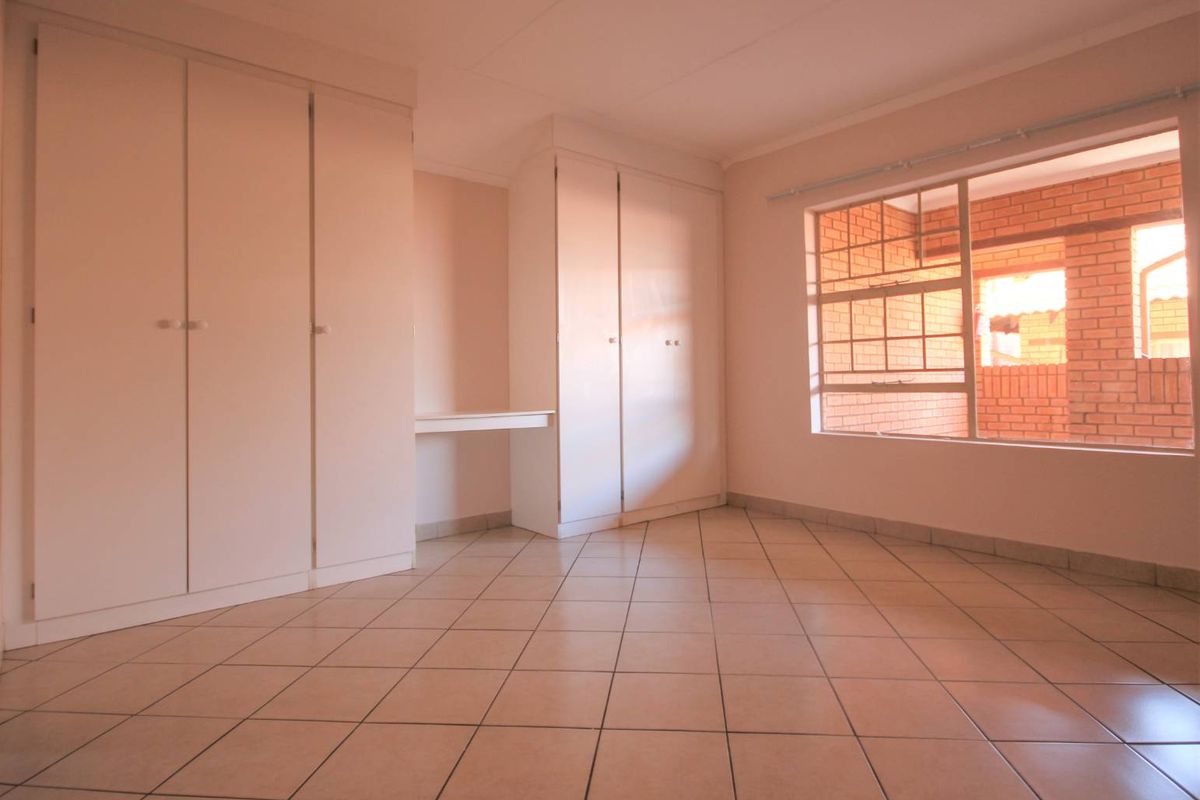 A bright, empty bedroom featuring built-in cupboards, a small integrated desk, tiled flooring, and a large window with security bars.