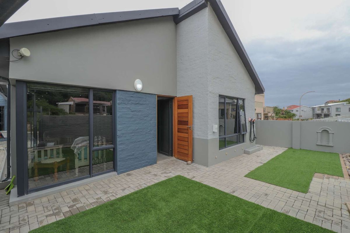 A modern exterior rear view of the house featuring a paved patio area, artificial lawn, and large glass sliding doors leading into the home.