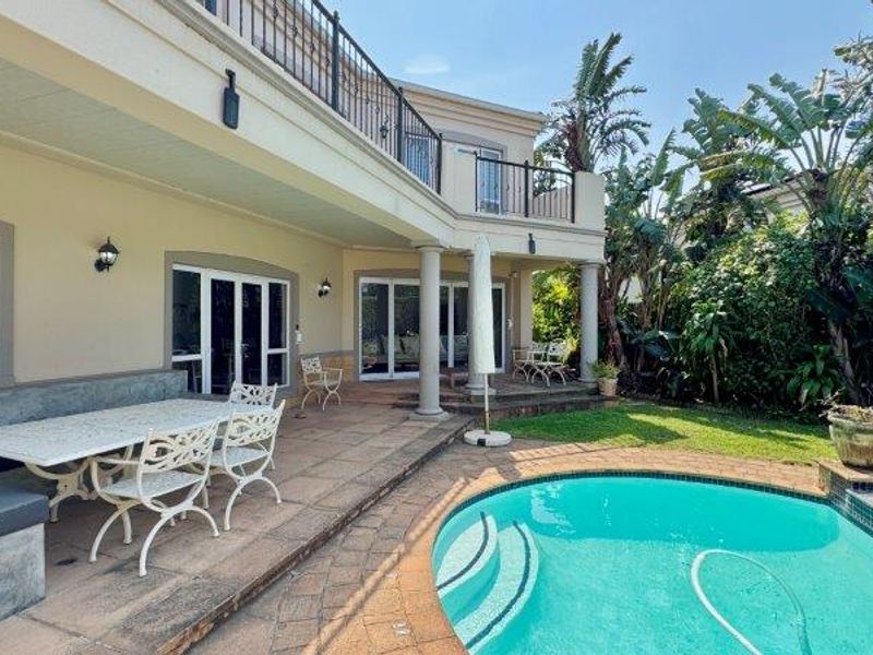 Spacious outdoor entertainment area featuring a covered patio, built-in concrete seating, and a swimming pool set within a landscaped garden.