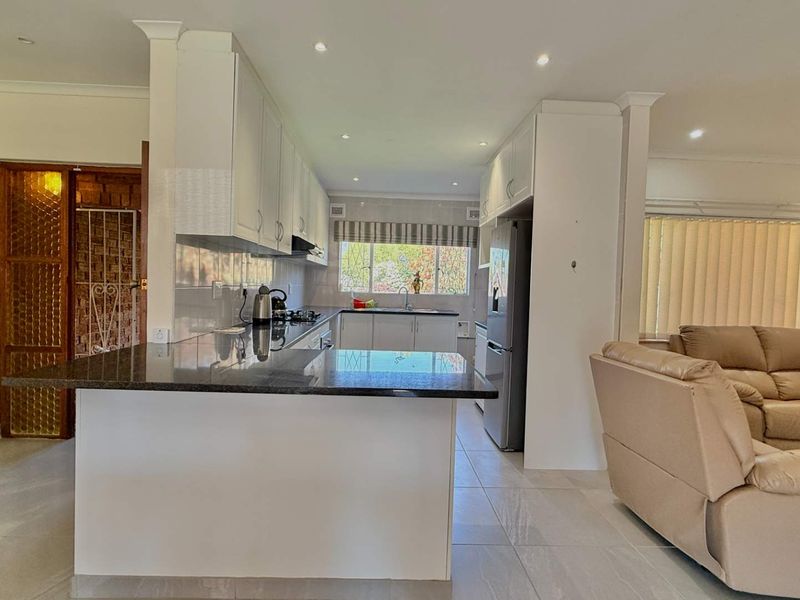 An open-plan kitchen featuring granite countertops, white cabinetry, and recessed lighting, seamlessly flowing into the adjacent living area with tiled flooring throughout.