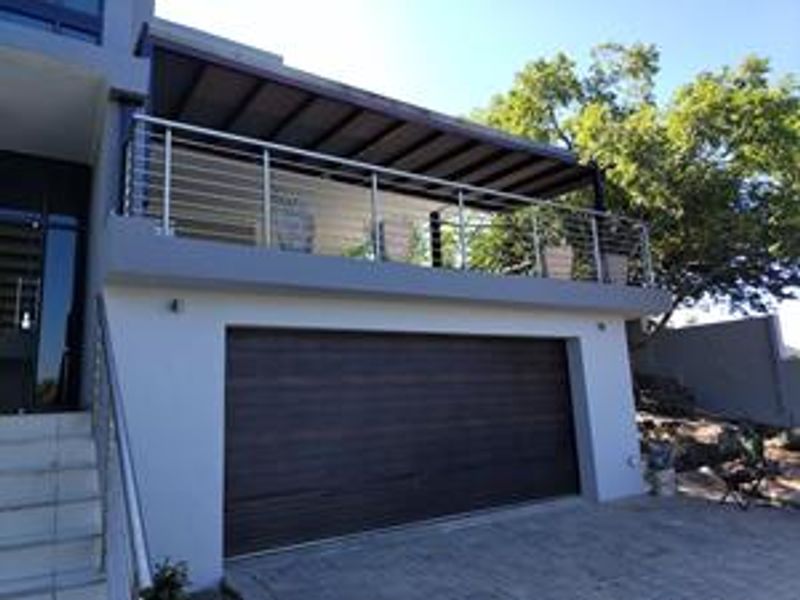 Front elevation featuring a modern garage door, a paved driveway, and an elevated balcony with stainless steel railings and a covered patio area.