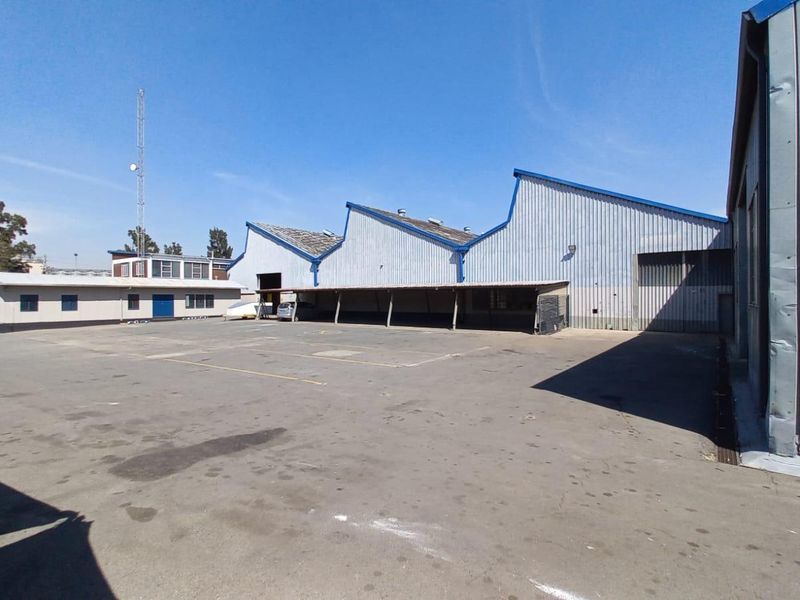 Rear industrial yard featuring a large asphalt area, covered loading bay, and sawtooth roof warehouse structure with metal cladding.