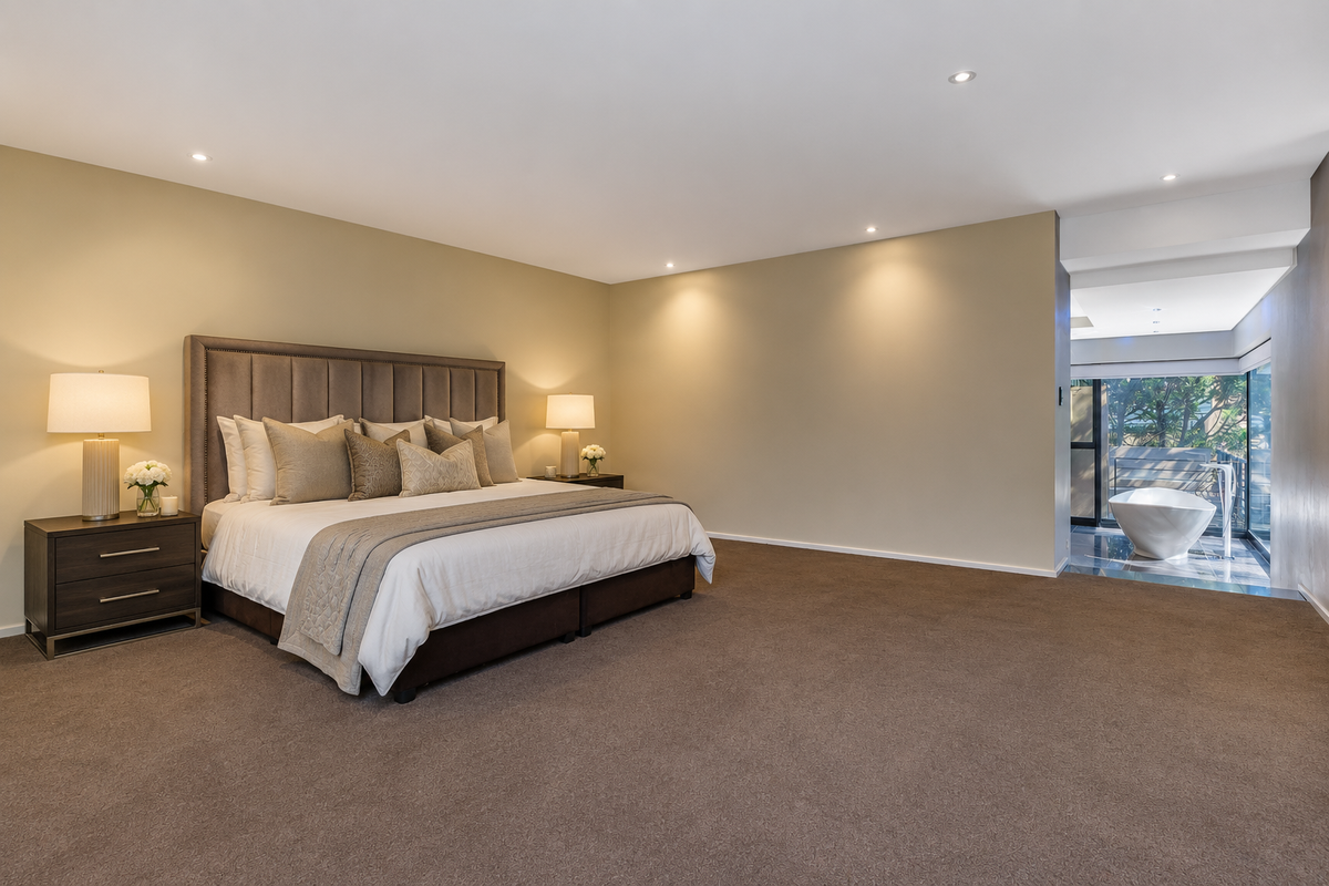Spacious master bedroom featuring neutral tones, plush carpeting, recessed lighting, and an open-plan ensuite area with a modern freestanding bathtub.