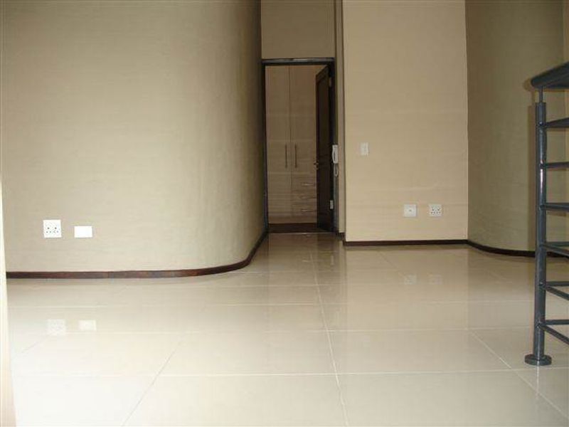 A spacious, empty hallway featuring neutral tiled flooring, curved wall architecture, and a metal balustrade leading to an adjacent room with built-in storage.