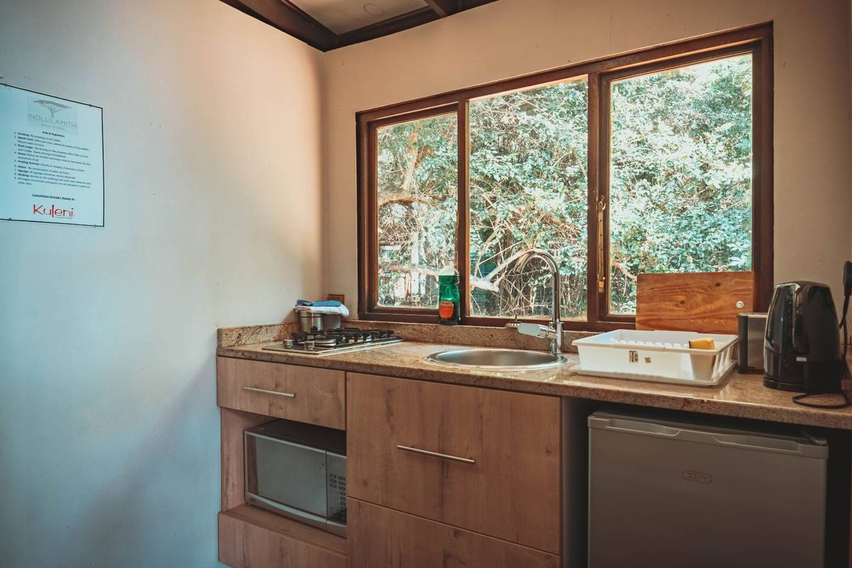 A compact kitchen featuring wood-finish cabinetry, granite countertops, a built-in gas hob, and a stainless steel sink overlooking a lush garden view.