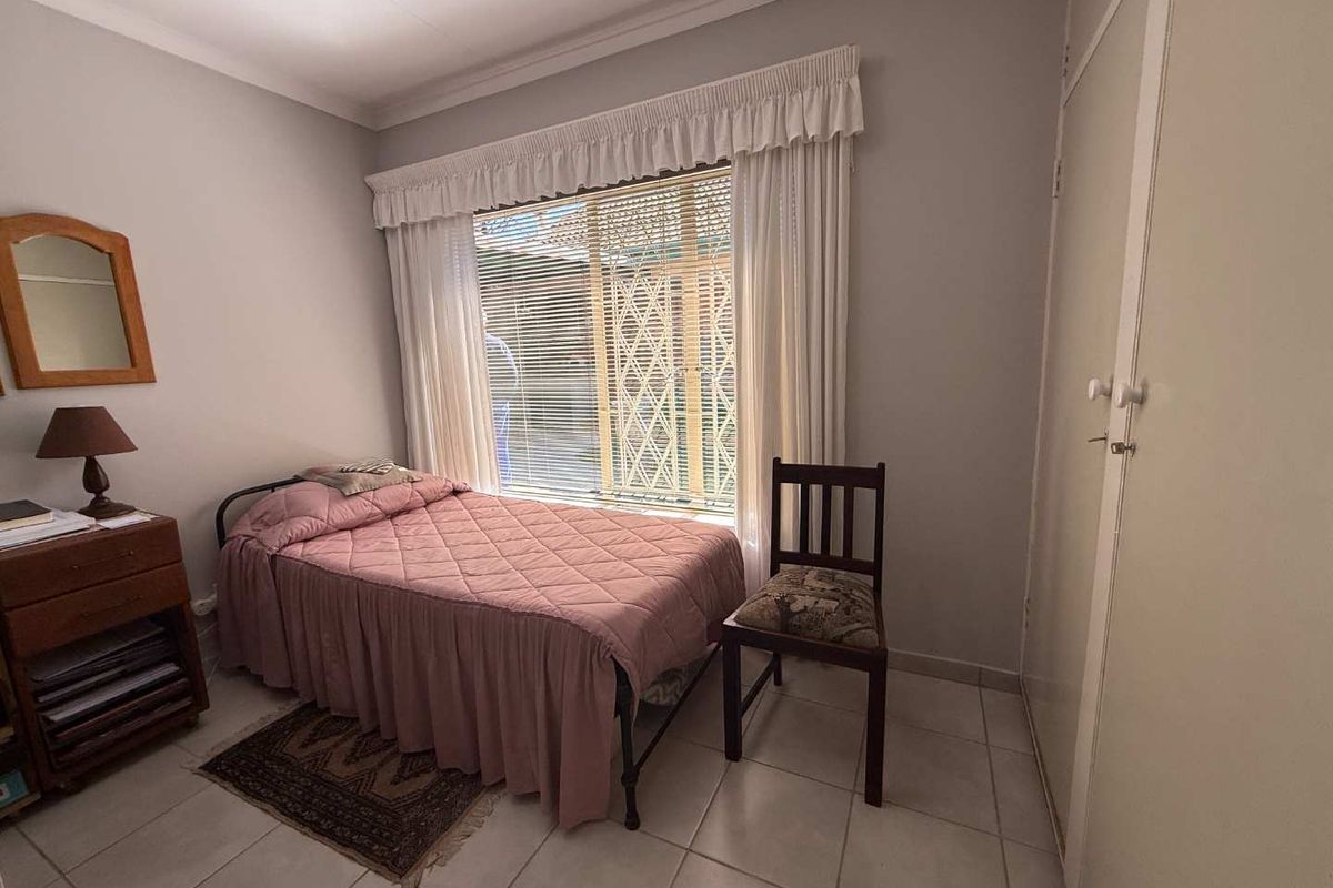 A functional bedroom featuring tiled flooring, a built-in cupboard, and a window fitted with security gates and blinds.