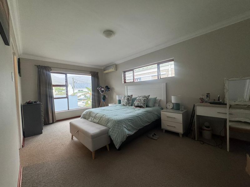 A spacious bedroom featuring neutral walls, carpeted flooring, and dual windows providing natural light, equipped with a wall-mounted air conditioning unit.