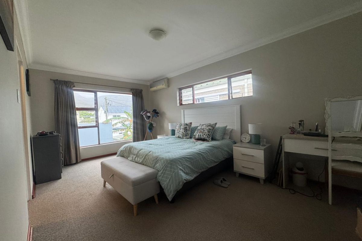 A spacious bedroom featuring neutral walls, carpeted flooring, and dual windows providing natural light, equipped with a wall-mounted air conditioning unit.