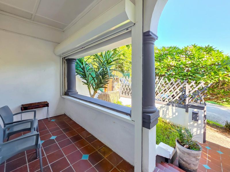 A covered patio area featuring tiled flooring, decorative columns, and a retractable security shutter, offering a view of the garden.
