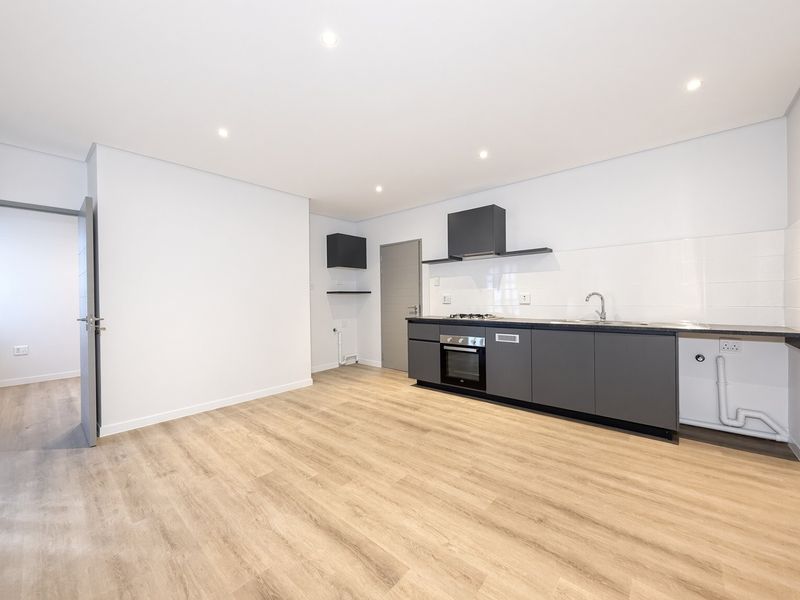 A modern, minimalist kitchen featuring dark grey cabinetry, built-in oven, gas hob, extractor fan, and light wood-look laminate flooring in an open-plan layout.