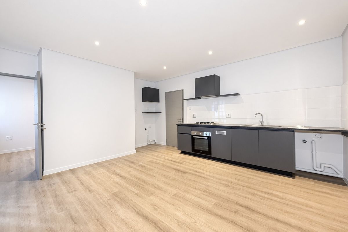 A modern, minimalist kitchen featuring dark grey cabinetry, built-in oven, gas hob, extractor fan, and light wood-look laminate flooring in an open-plan layout.