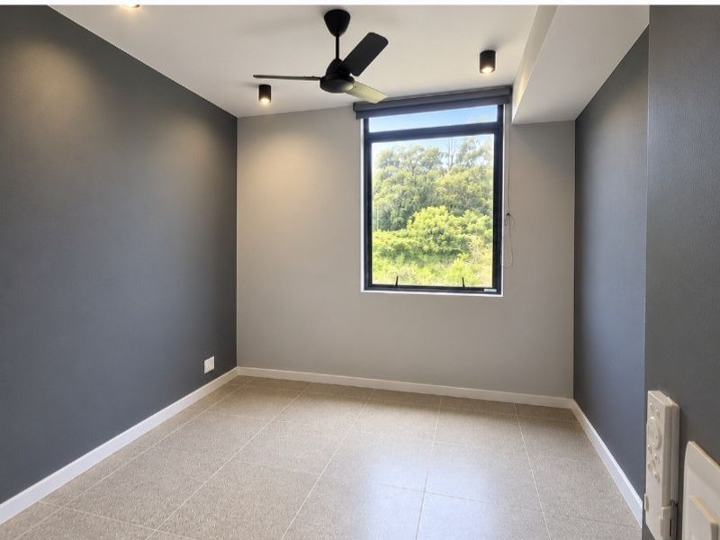 A bright, modern, and empty bedroom featuring neutral grey walls, tiled flooring, a ceiling fan, and a large window with a pleasant green view.