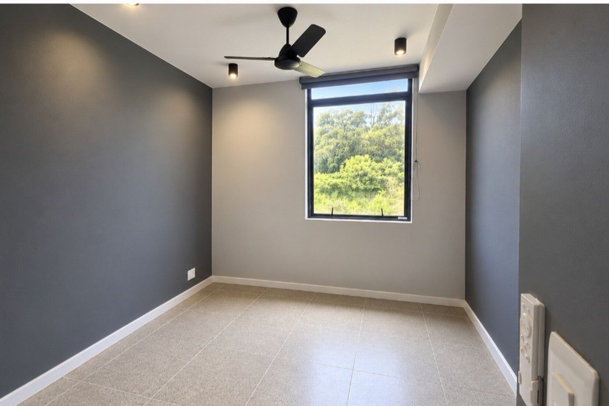 A bright, modern, and empty bedroom featuring neutral grey walls, tiled flooring, a ceiling fan, and a large window with a pleasant green view.