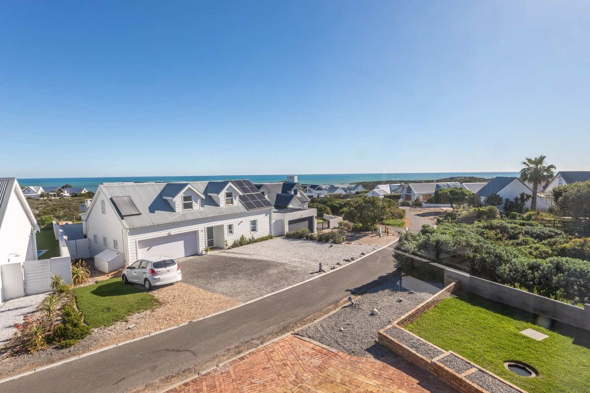 Aerial view of a coastal residential property featuring a white house with a metal roof, solar panels, and a paved driveway.