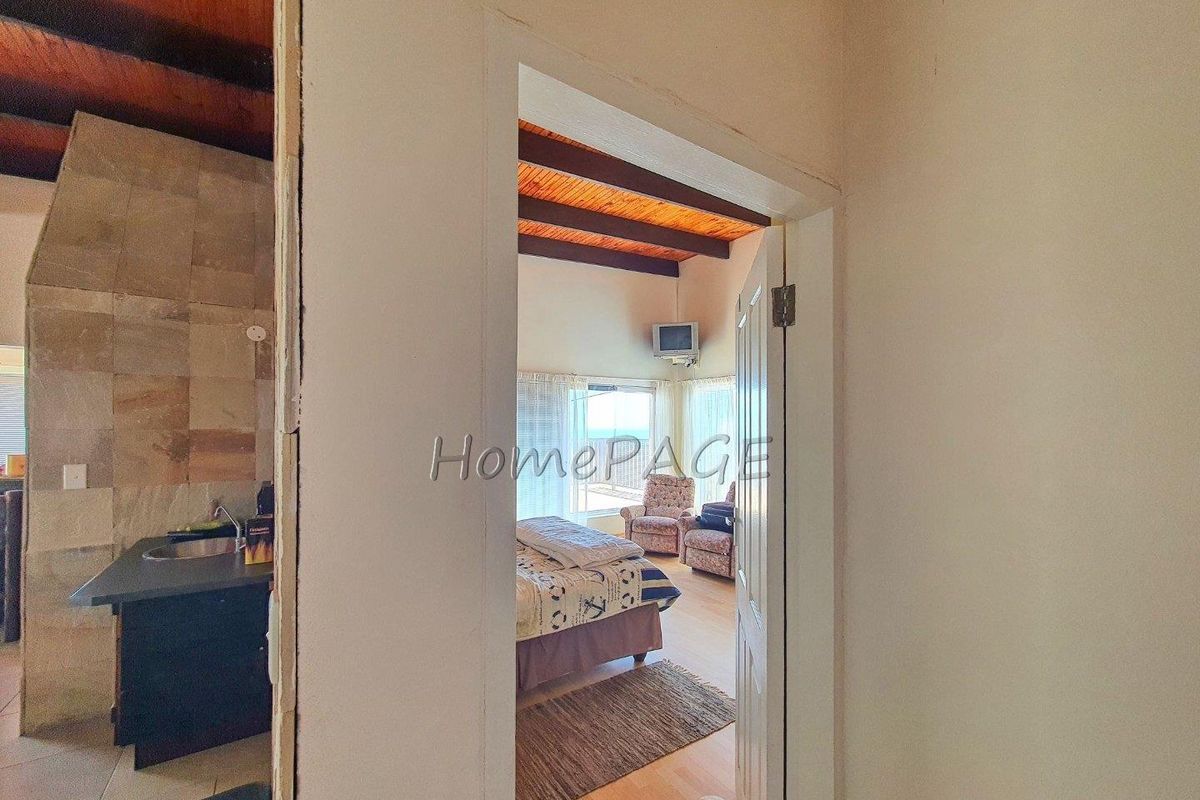 A view through a doorway into a bedroom featuring exposed timber ceiling beams and large windows, with a partial view of an adjacent kitchen area.