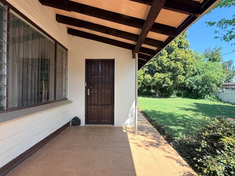 A covered patio area featuring tiled flooring, exposed timber rafters, and a security gate, overlooking a private garden.