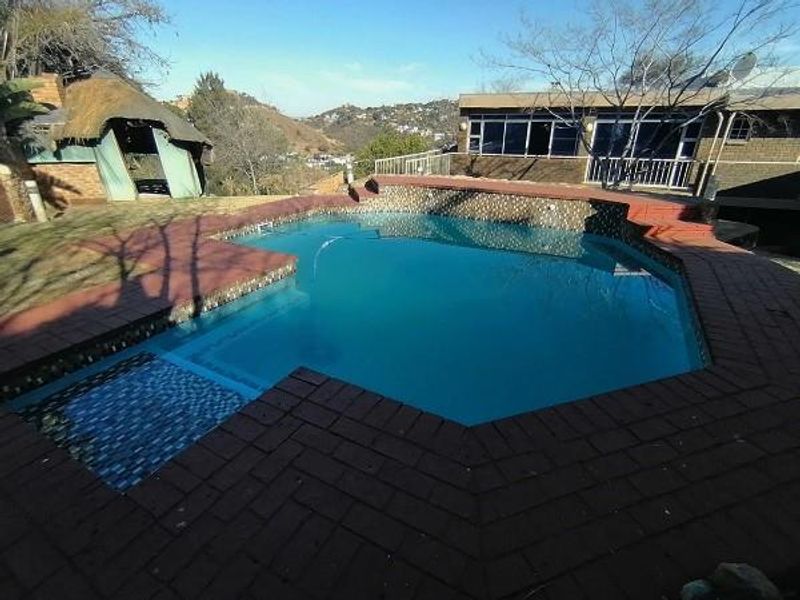 Outdoor pool area featuring brick paving, a raised retaining wall, and a nearby thatched lapa structure with scenic hillside views.