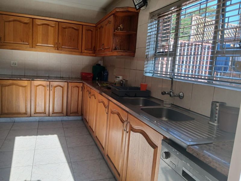 A functional kitchen featuring wooden cabinetry, double stainless steel sink, tiled splashbacks, and tiled flooring with natural light from the window.