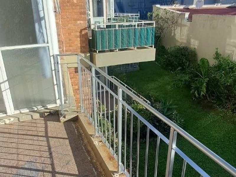 An outdoor balcony featuring a metal balustrade and tiled flooring, overlooking a communal garden area.