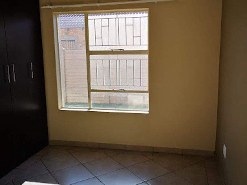 A clean, empty bedroom featuring tiled flooring, built-in cupboards, and a window fitted with security bars.