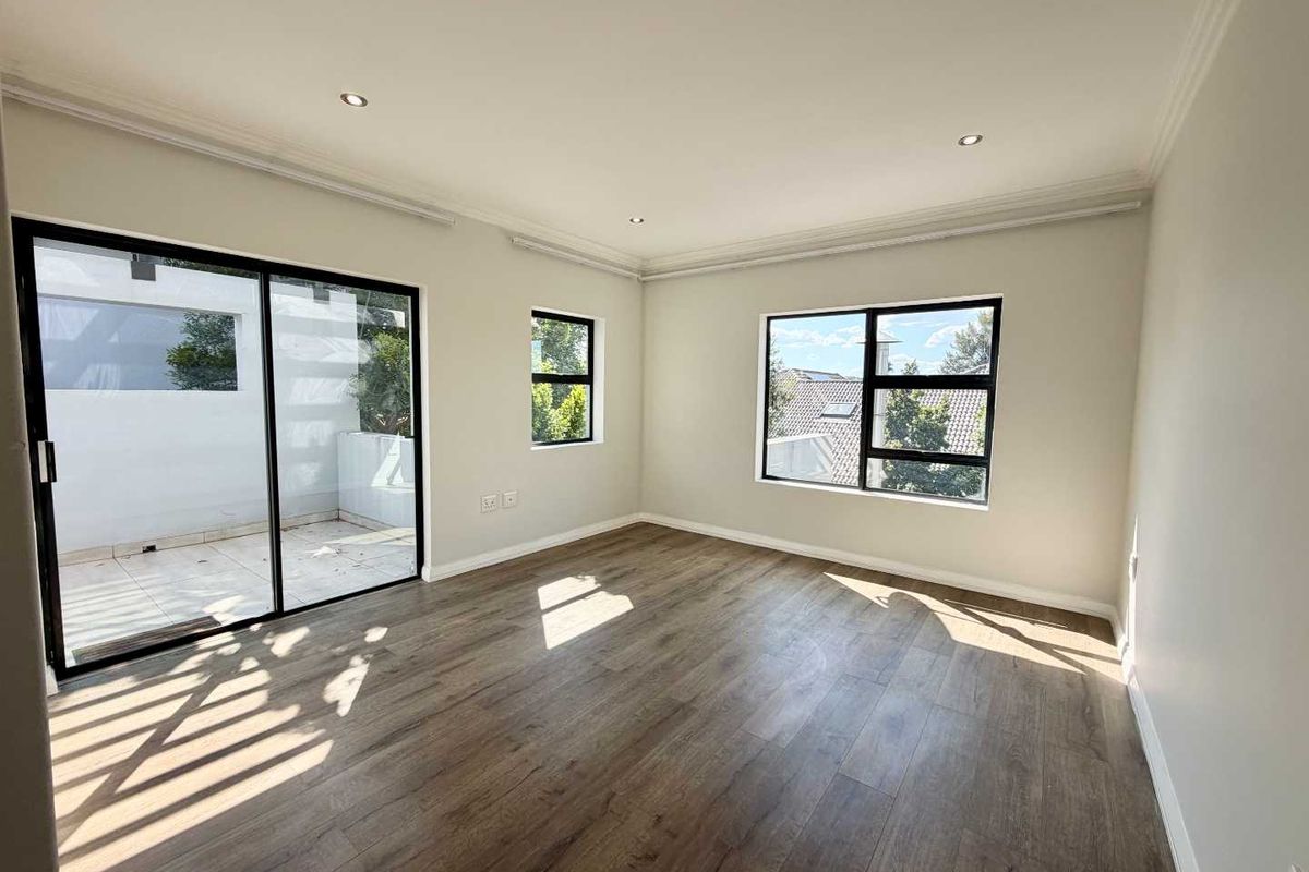 A bright, empty living room featuring modern laminate flooring, recessed downlights, and large windows with access to an outdoor patio area.