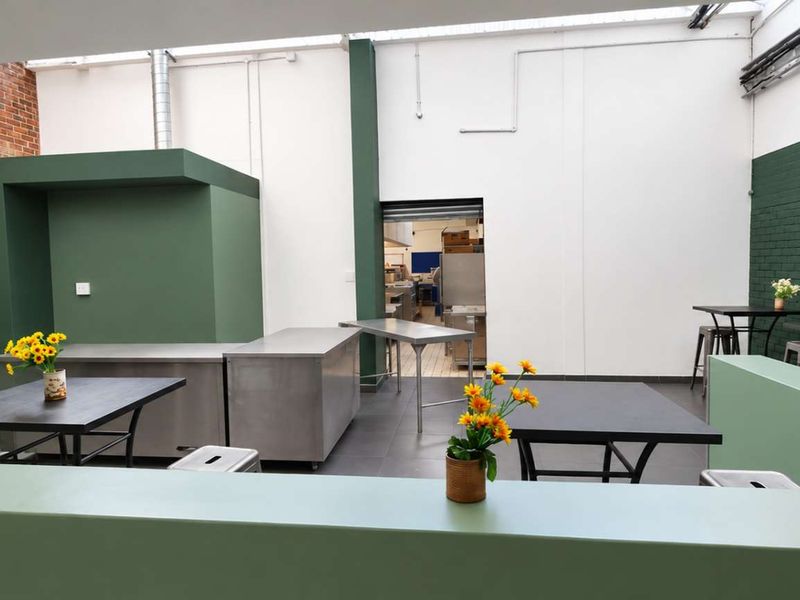Open-plan industrial space featuring exposed brickwork, painted accent walls, stainless steel prep surfaces, and natural light from overhead skylights.