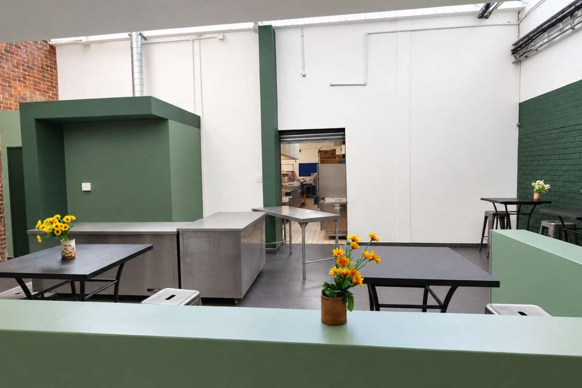 Open-plan industrial space featuring exposed brickwork, painted accent walls, stainless steel prep surfaces, and natural light from overhead skylights.