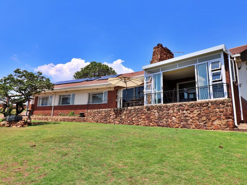 Rear view of a house featuring a stone-clad raised patio, brick and plaster facade, tiled roof with solar panels, and a prominent stone chimney.