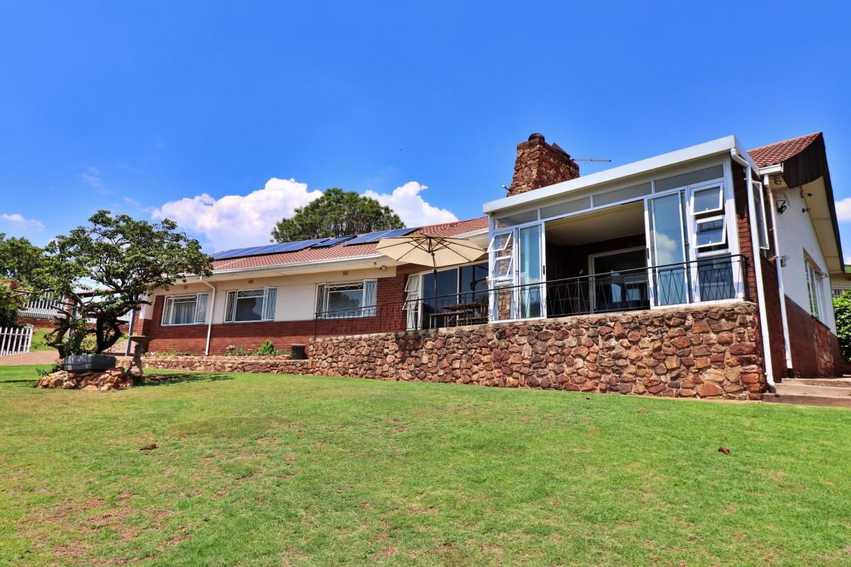 Rear view of a house featuring a stone-clad raised patio, brick and plaster facade, tiled roof with solar panels, and a prominent stone chimney.