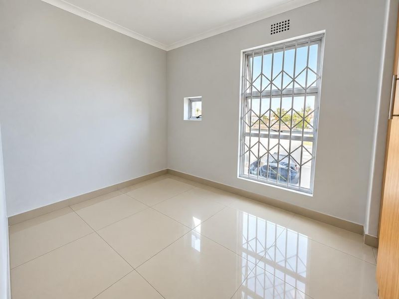 A bright, empty bedroom featuring neutral tiled flooring, built-in cupboard storage, and windows fitted with security bars.