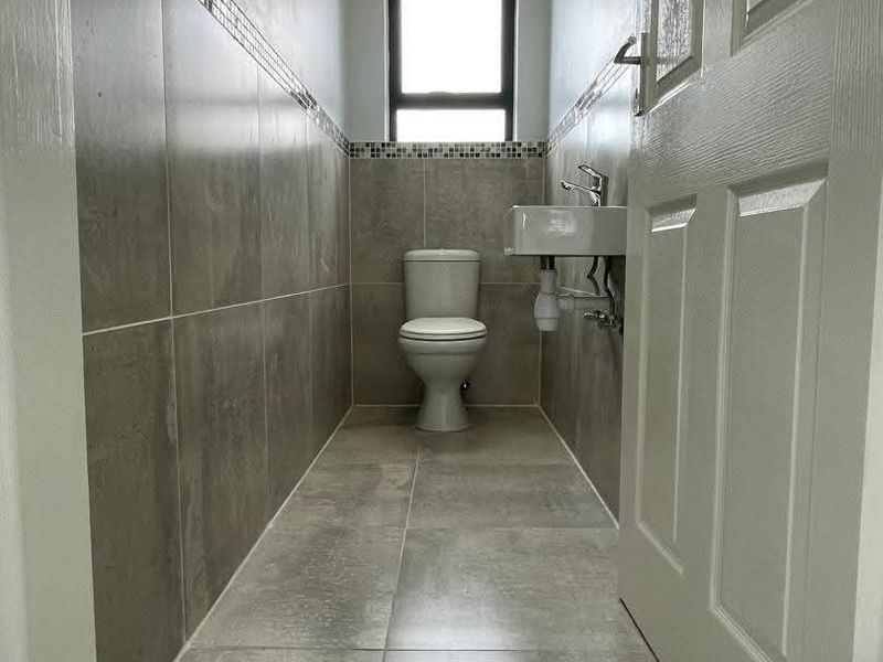 A clean, modern guest bathroom featuring neutral grey floor and wall tiles, a wall-mounted basin, and a toilet.