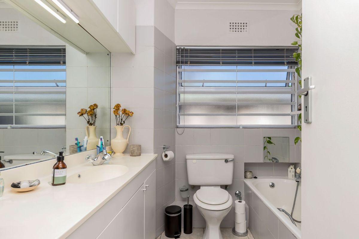 A functional bathroom featuring a vanity with integrated sink, large wall mirror, toilet, and a bathtub with shower mixer, finished with neutral wall tiles.