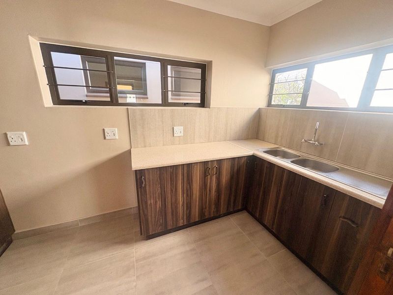 A clean, empty kitchen area featuring wood-look laminate cabinetry, a double stainless steel sink, tiled splashbacks, and large windows with security bars.