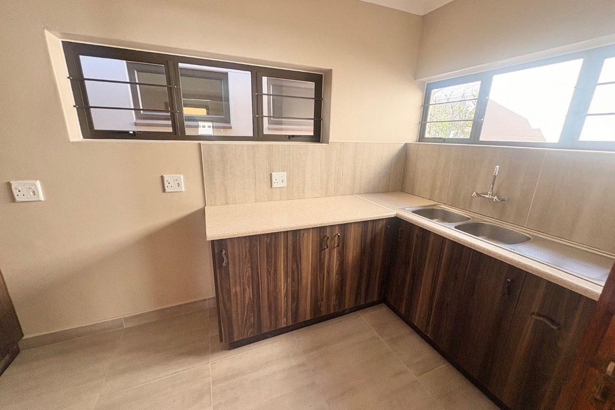 A clean, empty kitchen area featuring wood-look laminate cabinetry, a double stainless steel sink, tiled splashbacks, and large windows with security bars.