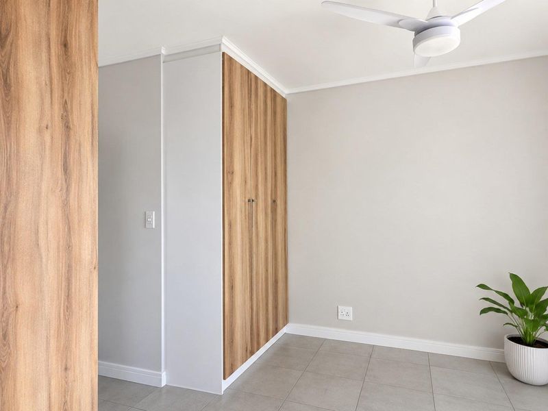 A bright, modern bedroom featuring neutral walls, tiled flooring, a ceiling fan, and sleek wood-finish built-in cupboards.