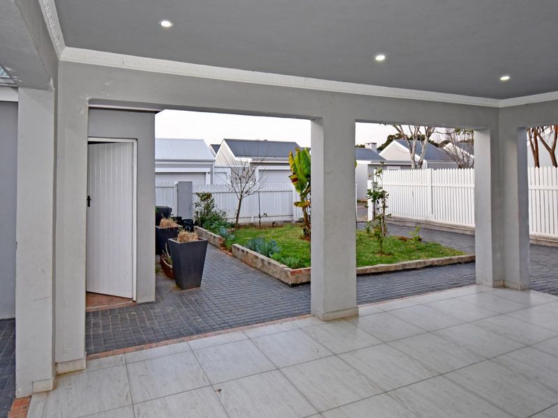 A covered patio area featuring tiled flooring and recessed lighting, overlooking a neat paved courtyard with a raised garden bed and white fencing.