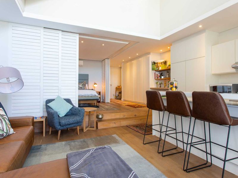 Modern open-plan apartment featuring a sleek kitchen with island, timber flooring, and a raised bedroom area separated by white sliding shutters.