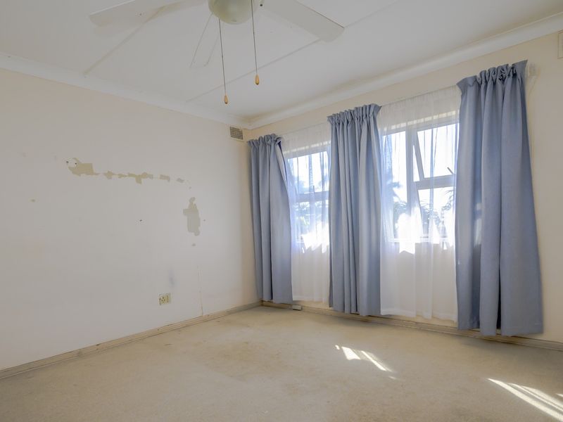 An empty bedroom featuring a ceiling fan, large windows with curtains, and carpet flooring. The room requires cosmetic updates to the walls and flooring.