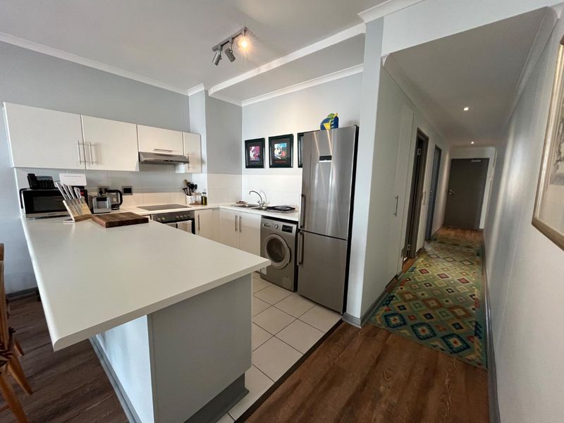 Open-plan kitchen featuring white cabinetry, integrated appliances, and a breakfast bar, seamlessly transitioning into a hallway with wood-look flooring.