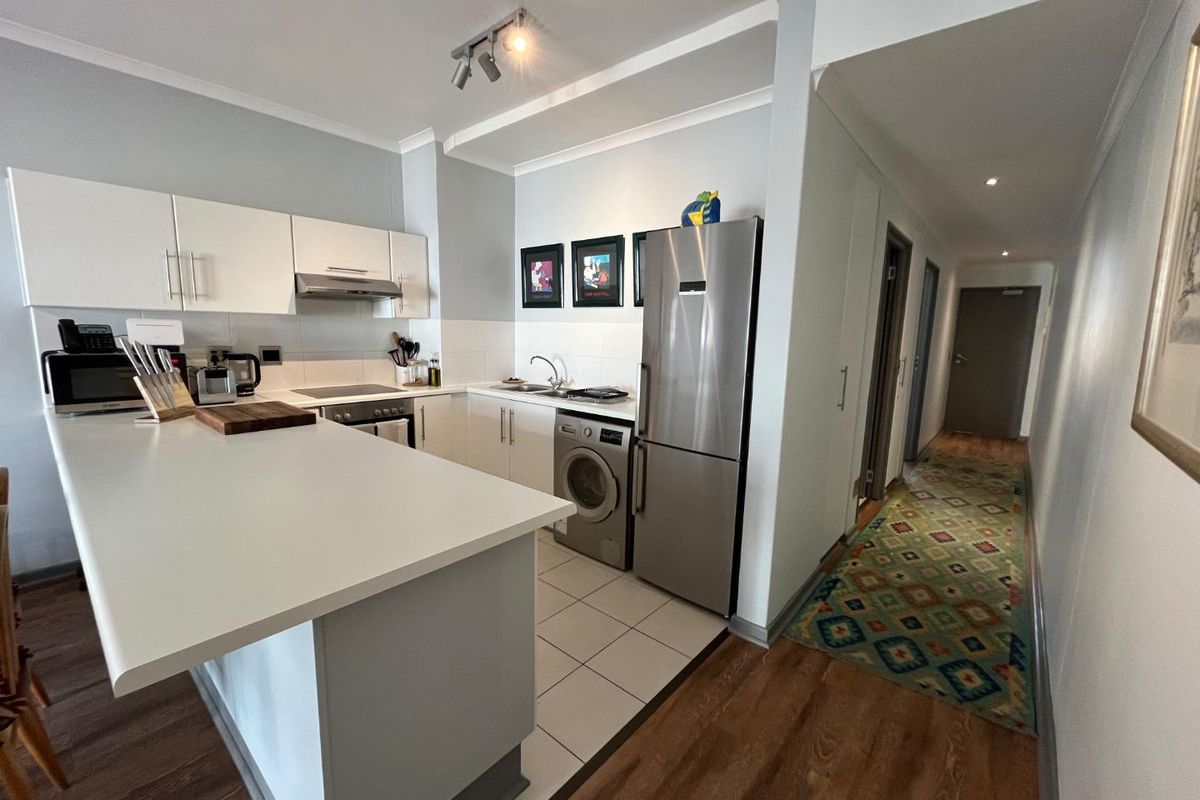 Open-plan kitchen featuring white cabinetry, integrated appliances, and a breakfast bar, seamlessly transitioning into a hallway with wood-look flooring.