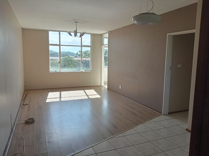 An empty, sunlit living room featuring a combination of laminate and tiled flooring, large windows, and neutral wall tones.