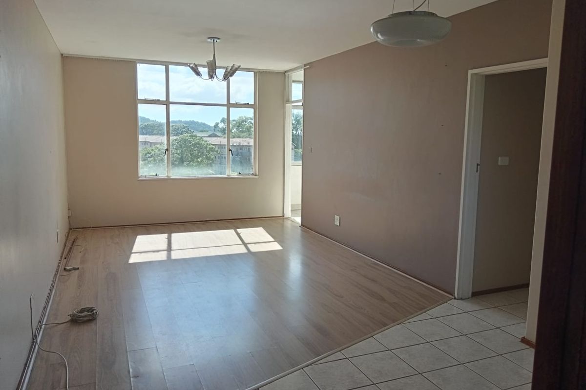 An empty, sunlit living room featuring a combination of laminate and tiled flooring, large windows, and neutral wall tones.