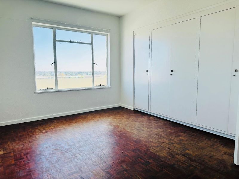 An empty bedroom featuring original parquet flooring, built-in storage cupboards, and a large window offering natural light and exterior views.