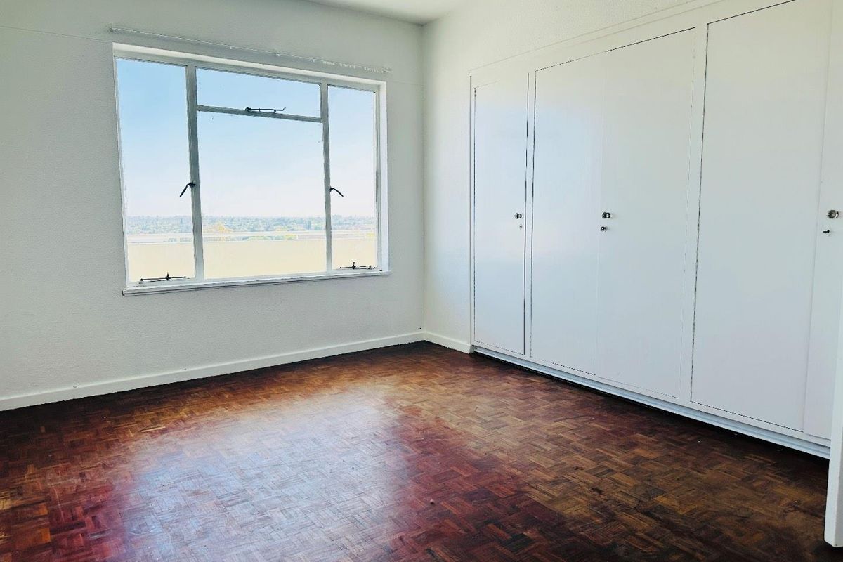 An empty bedroom featuring original parquet flooring, built-in storage cupboards, and a large window offering natural light and exterior views.