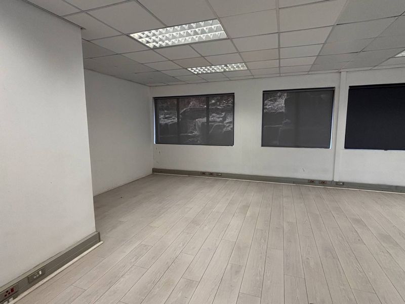 A bright, empty office space featuring laminate flooring, suspended ceilings with recessed lighting, and large windows equipped with roller blinds.