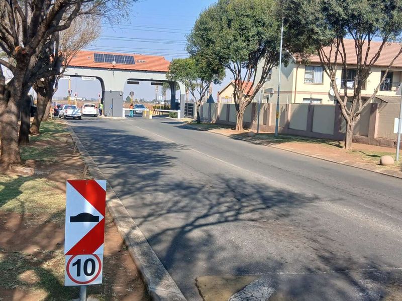 View of the residential complex entrance featuring a security gatehouse, perimeter walling, and paved access road.