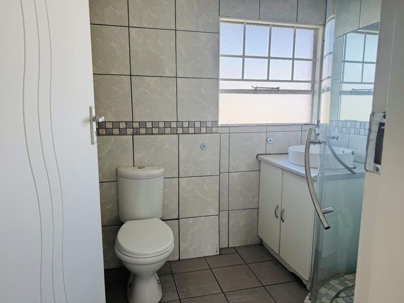 A functional bathroom featuring a toilet, vanity with vessel sink, and a glass-enclosed shower, finished with neutral wall and floor tiling.