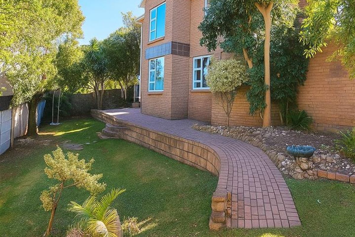 A well-maintained rear garden featuring a brick-paved pathway, tiered retaining walls, and established landscaping adjacent to the two-story brick house.