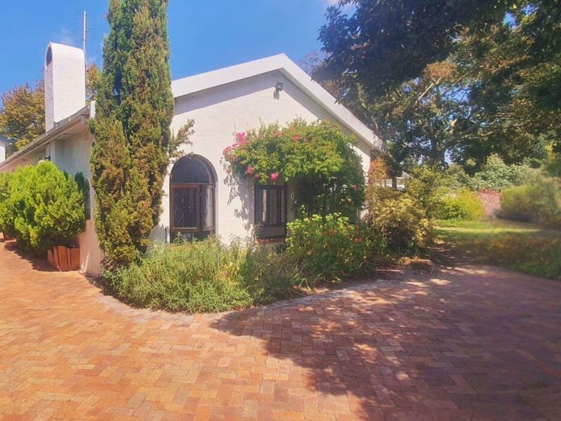 A charming cottage-style home featuring a brick-paved driveway, arched windows, and a prominent chimney, set within a lush, landscaped garden.